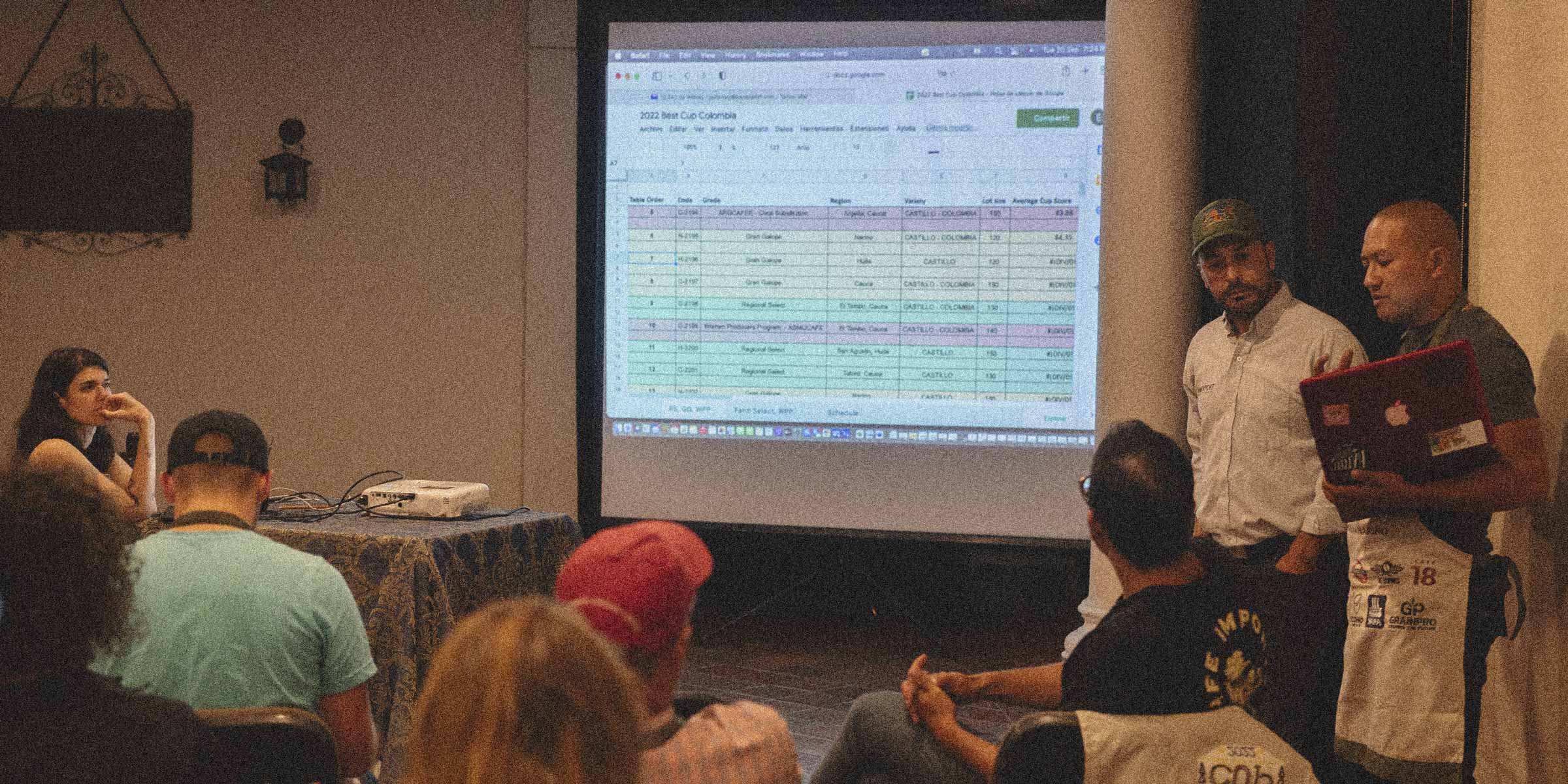Person giving a presentation in front of an audience with a projector screen displaying data about a coffee competition