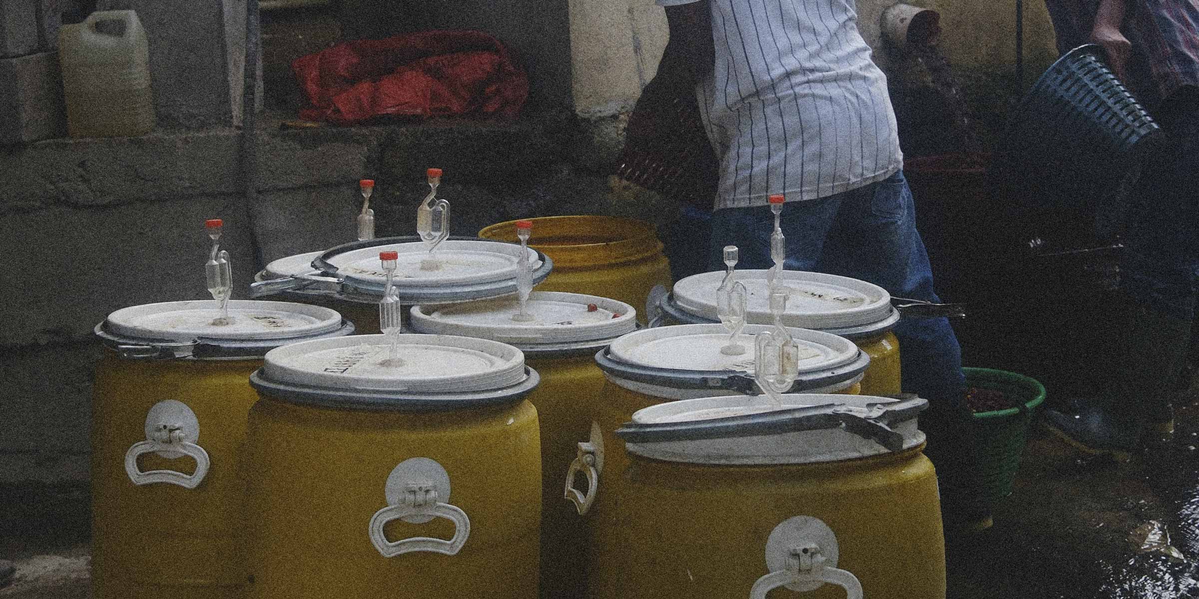 Yellow barrels with white lids on a stone floor, with people and bottles in the background.