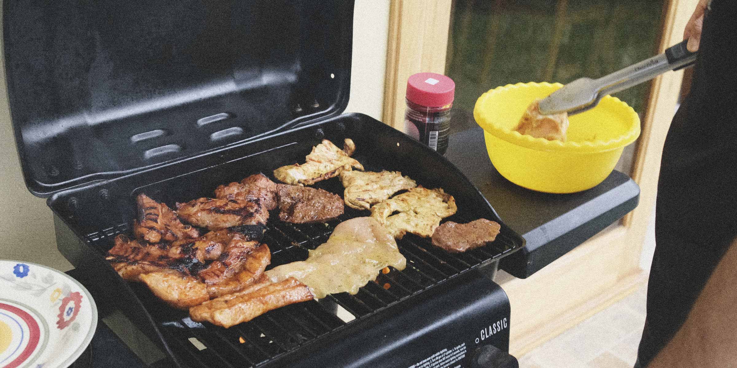 Grill with food being cooked and a person using tongs, with a yellow bowl and spices in the background.