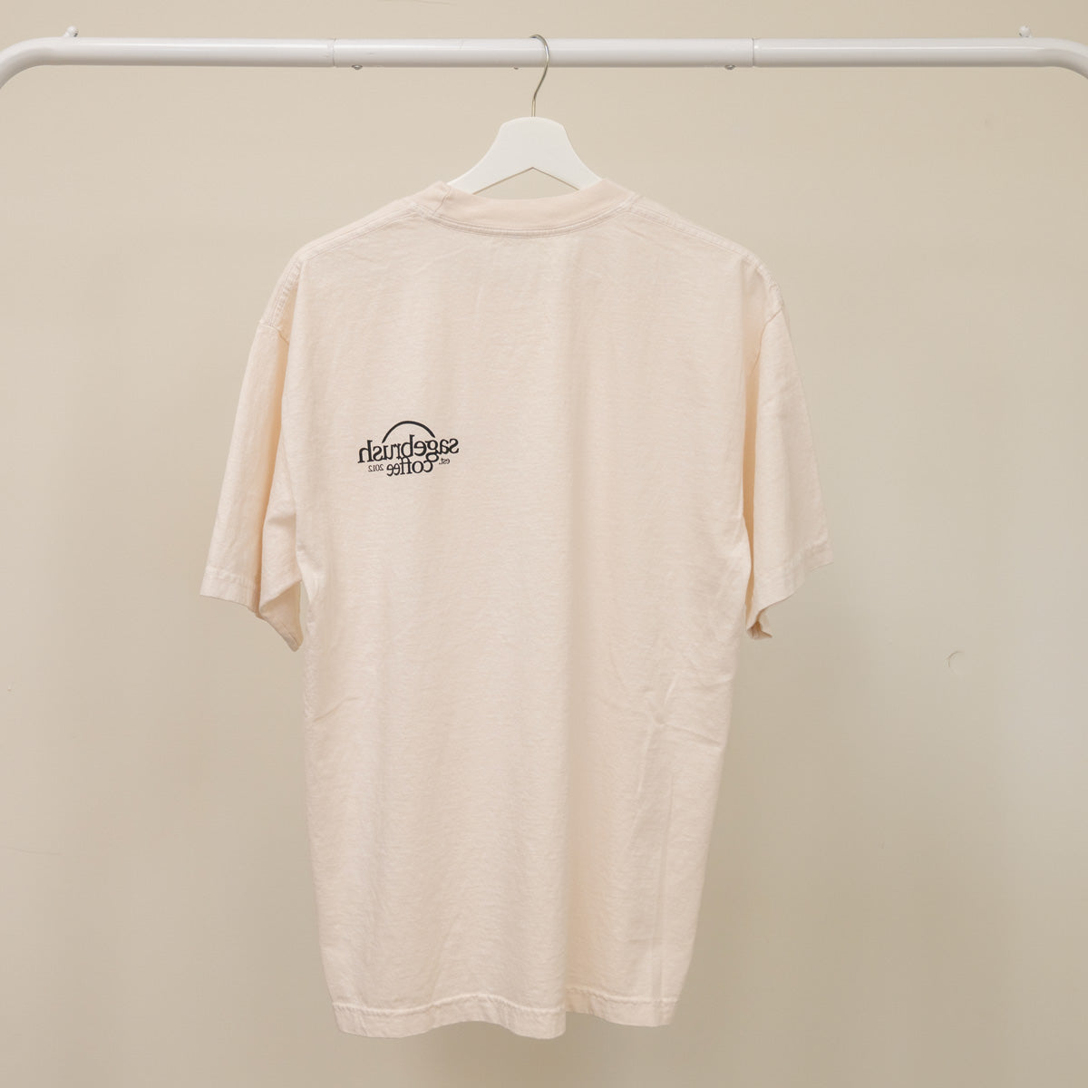 Beige t-shirt with a logo on a hanger against a beige wall