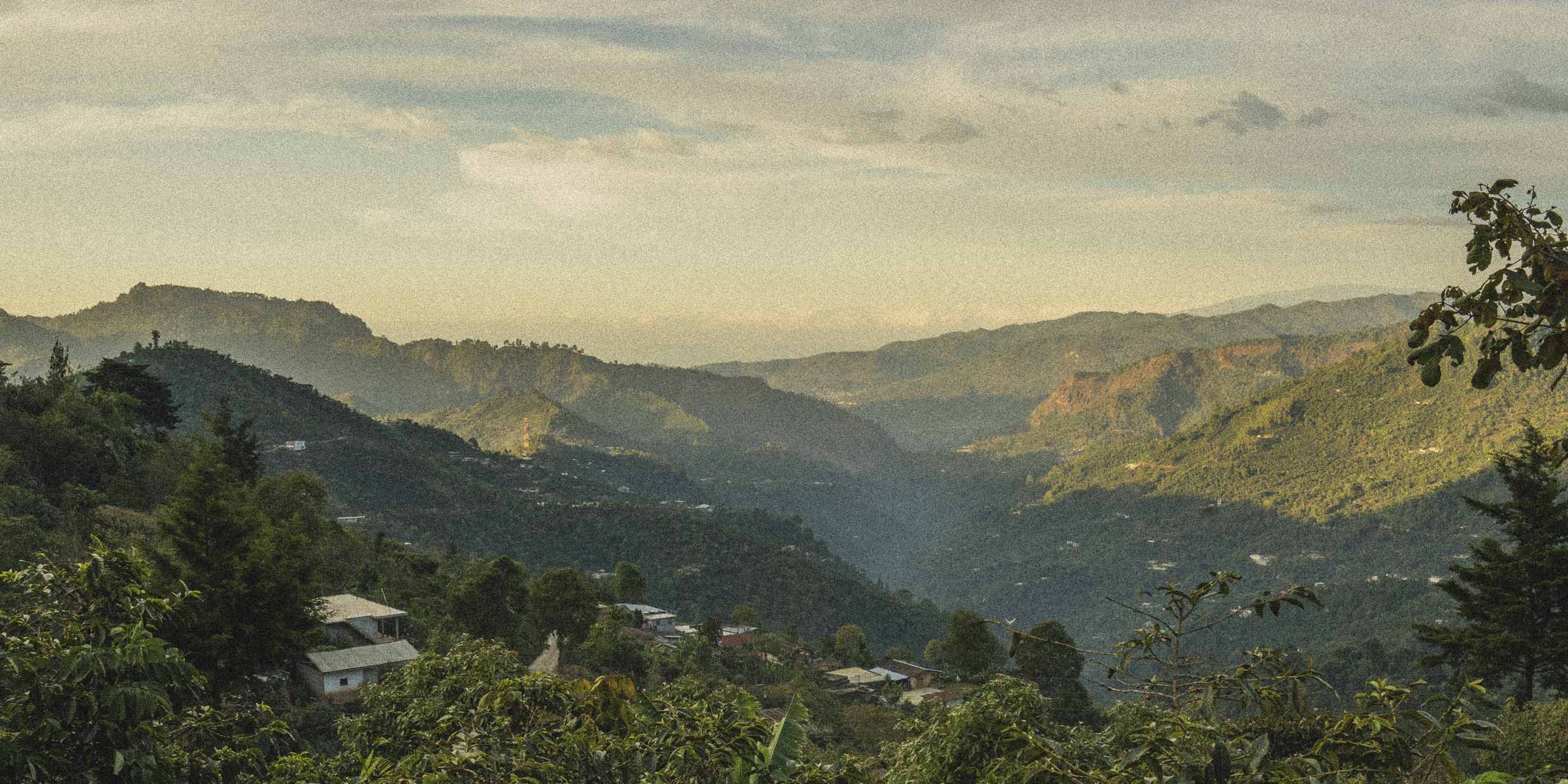 Hilly landscape of the mountains of Huehuetanango with greenery and a few buildings, under a cloudy sky.