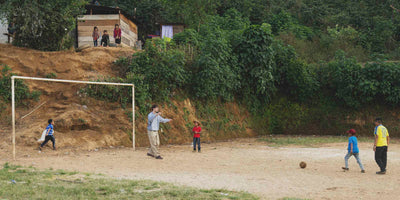 Children playing soccer on a dirt field with a goalpost, surrounded by greenery.