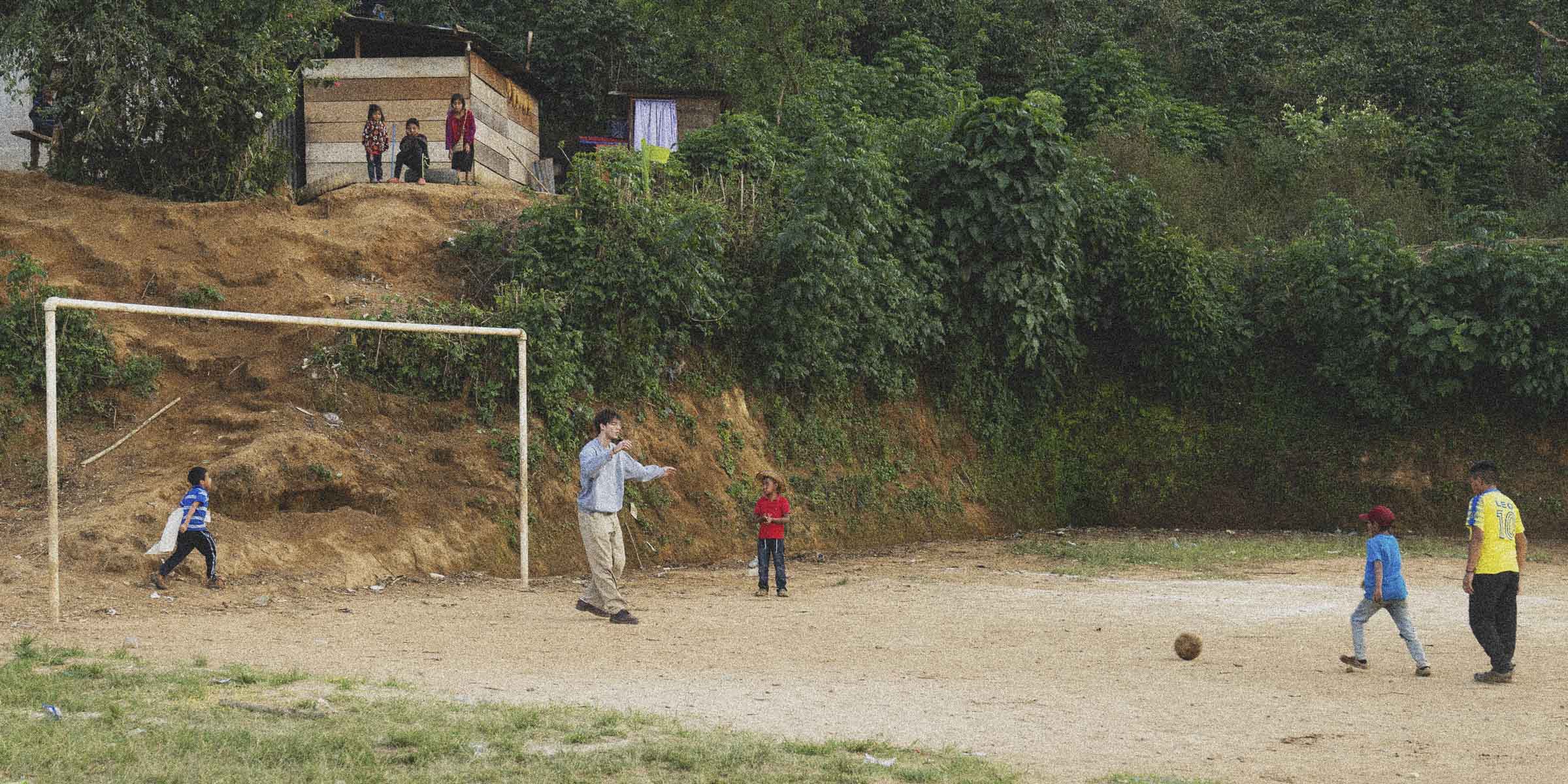 Children playing soccer on a dirt field with a goalpost, surrounded by greenery.