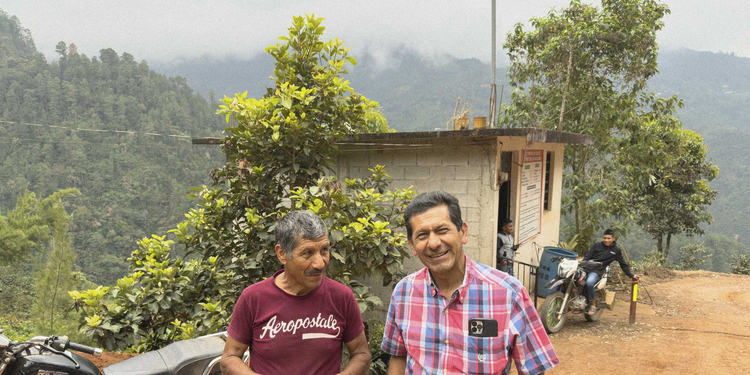 Villatoro brothers standing on a dirt road with a small building and motorcycle in the background, surrounded by greenery.