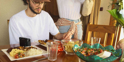 Person sitting at a table with food and drinks, another person standing in the background.