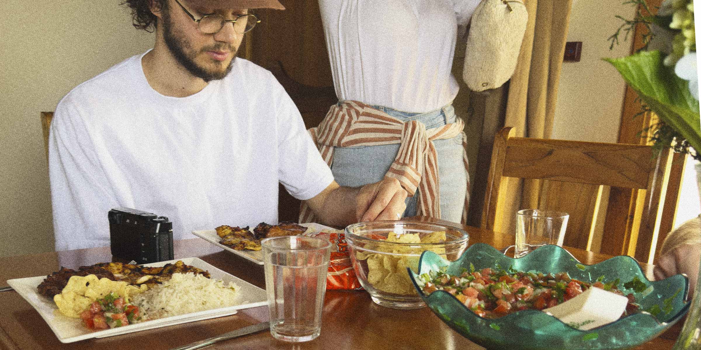 Person sitting at a table with food and drinks, another person standing in the background.