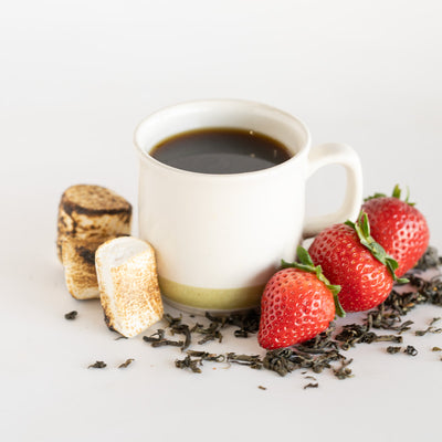 White mug with black coffee, strawberries, toasted marshmallow, and tea leaves on a light background