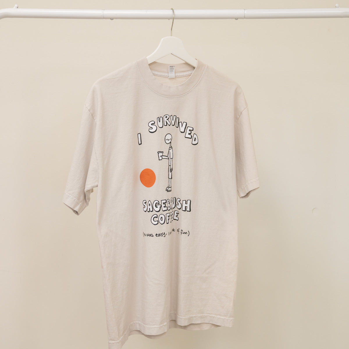 Beige t-shirt with graphic design on a hanger against a beige wall