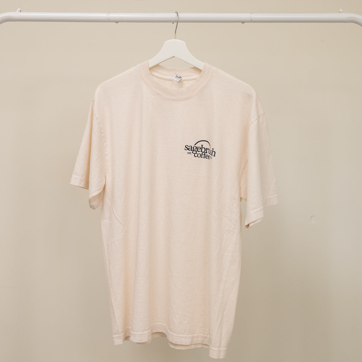 Beige t-shirt with a sagebrush coffee logo on a hanger against a beige wall