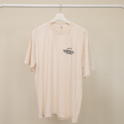 Beige t-shirt with a sagebrush coffee logo on a hanger against a beige wall